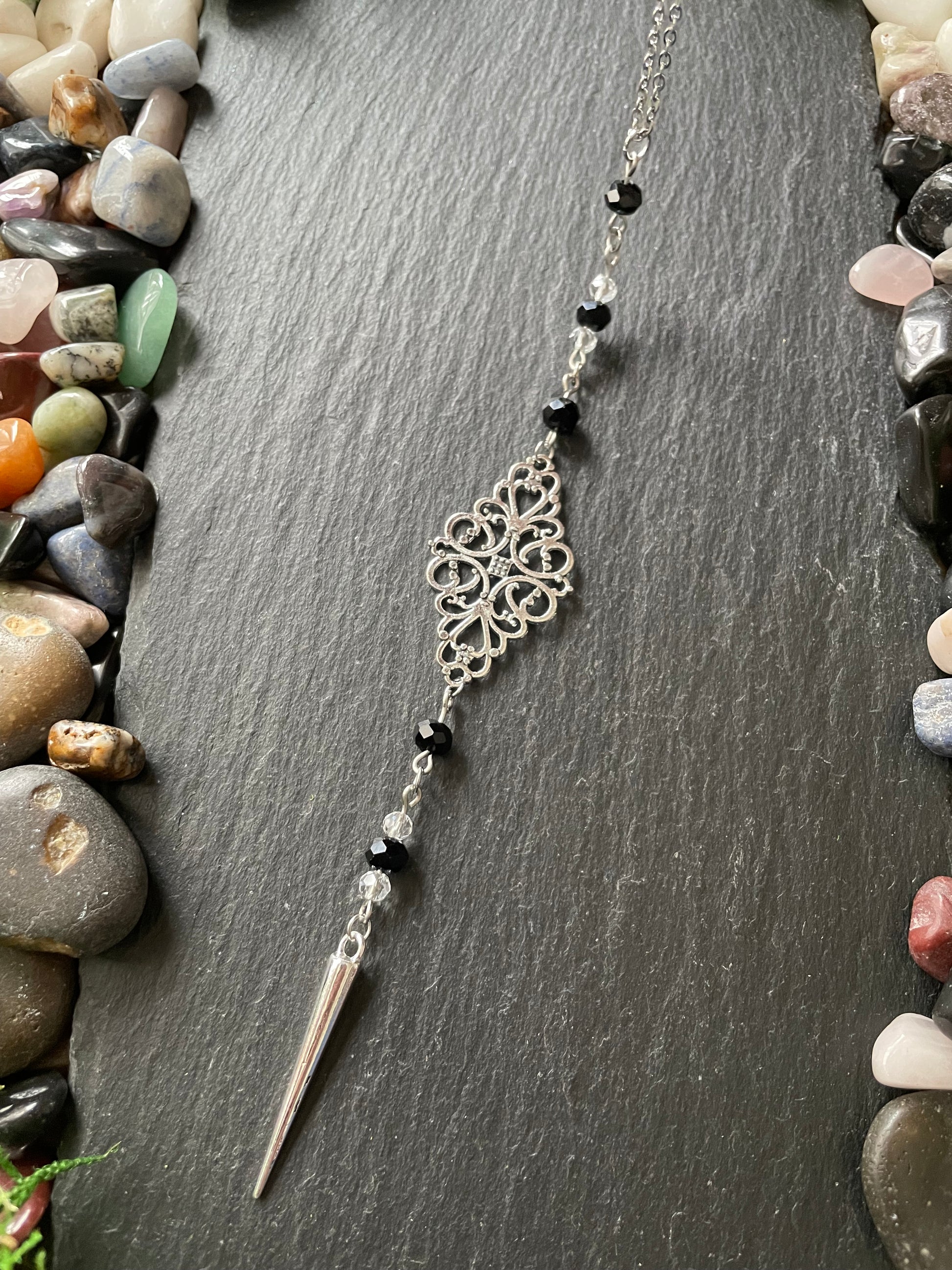 Silver car mirror charm with black beads on a dark stone surface with pebbles around
