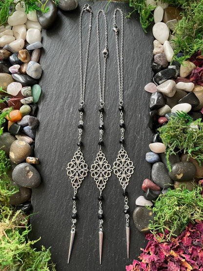 Two silver car mirror charms with decorative pendants on a dark slate surface surrounded by stones and plants.