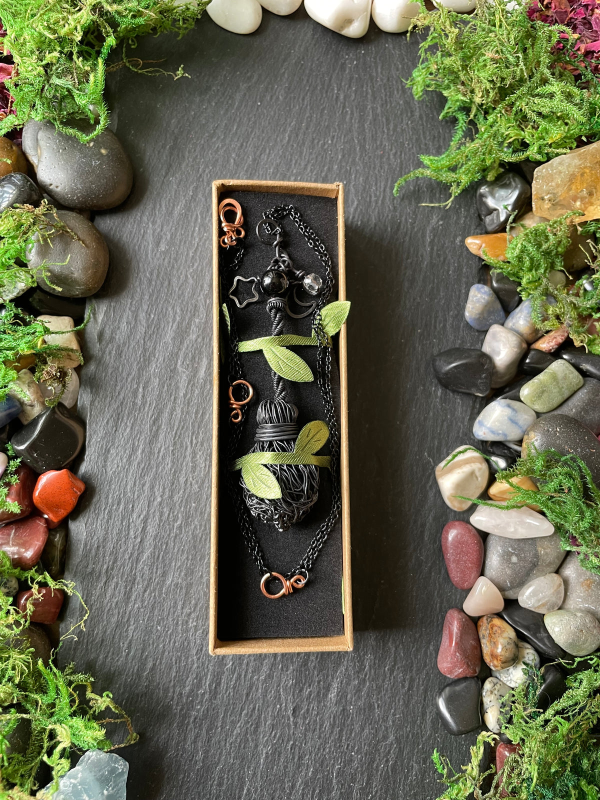 A jewelry box containing a black metal pendant necklace and matching chain, styled with moss and small green leaves on a dark slate surface surrounded by stones.