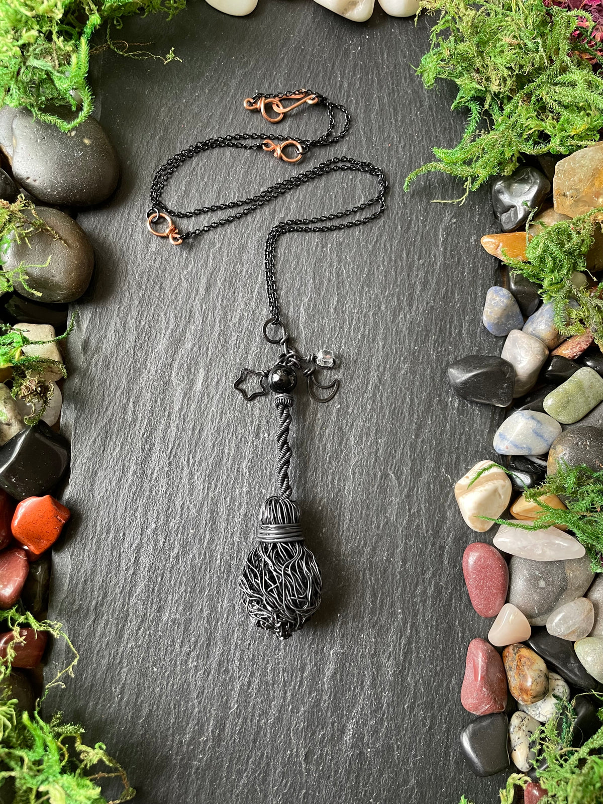 A handmade black metal pendant necklace, displayed on a dark slate background surrounded by decorative moss and stones.