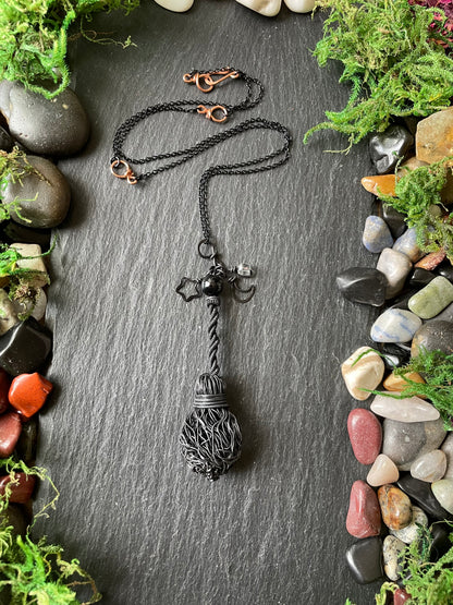 A handmade black metal pendant necklace, displayed on a dark slate background surrounded by decorative moss and stones.