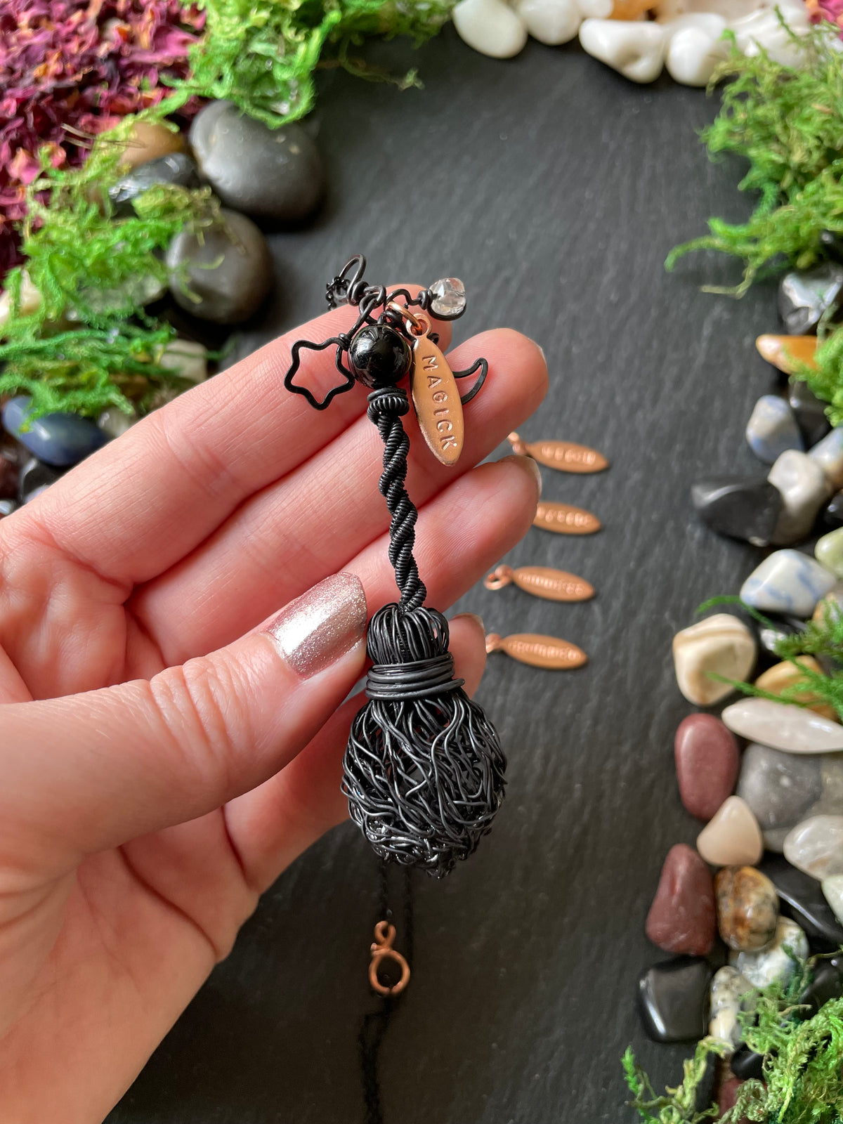 A hand holding a black twisted broom pendant with a small black bead. The background is a dark slate surface decorated with moss and smooth stones.