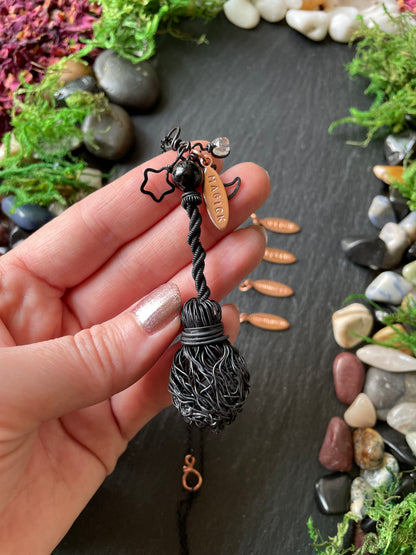 A hand holding a black twisted broom pendant with a small black bead. The background is a dark slate surface decorated with moss and smooth stones.