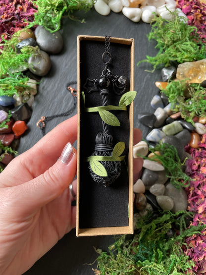 A hand holds a rectangular jewelry box containing a black metal broom pendant against a dark slate background decorated with moss and stones.
