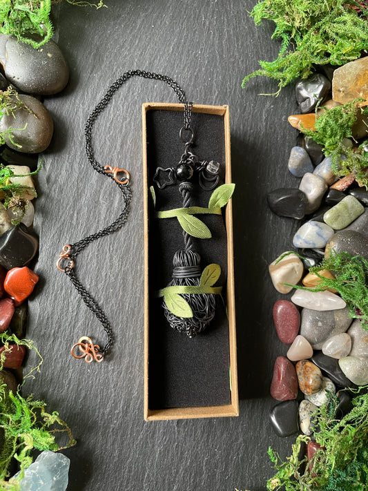 A black pendant necklace displayed in a rectangular wooden box on a dark slate background surrounded by stones and moss.