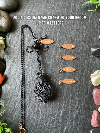 A black broom pendant, displayed on a dark slate background with decorative stones and text reading 'ADD A CUSTOM NAME CHARM TO YOUR BROOM UP TO 8 LETTERS.' Four small copper-colored name charms are shown beside it.