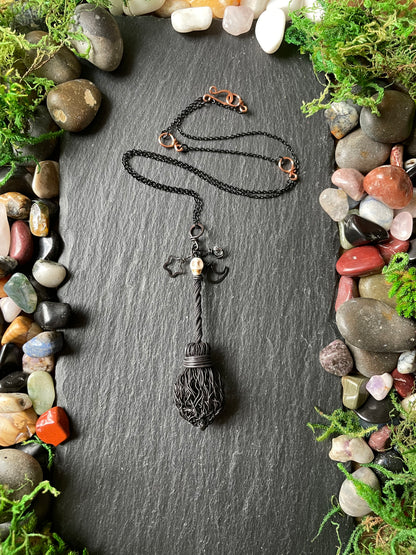 A black broom necklace with star and moon charms, arranged on a dark slate surface surrounded by smooth stones and green moss.