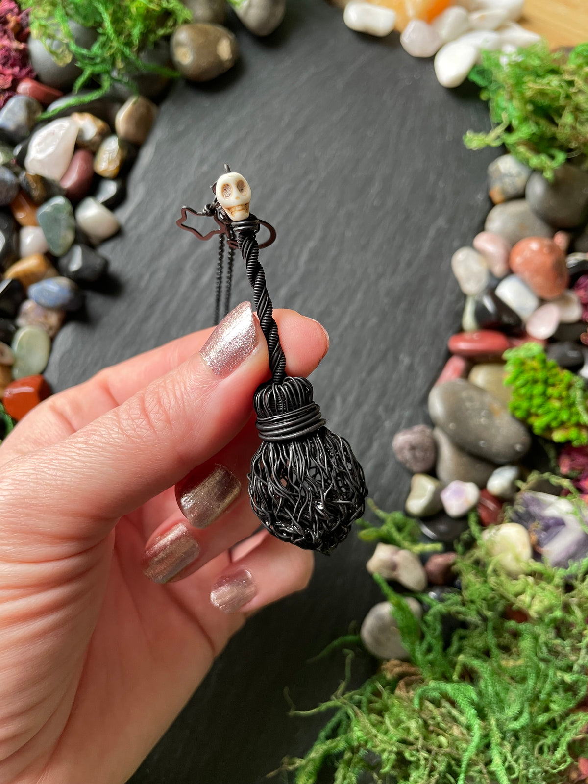 A hand with glittery nails holds a small black wire-wrapped broom charm with a tiny skull at the top, set against a dark slate background surrounded by decorative stones and green moss.