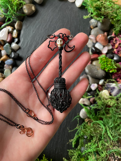 A hand holding a black witch-style broom pendant necklace with a black chain, surrounded by green moss and small stones on a dark slate background.