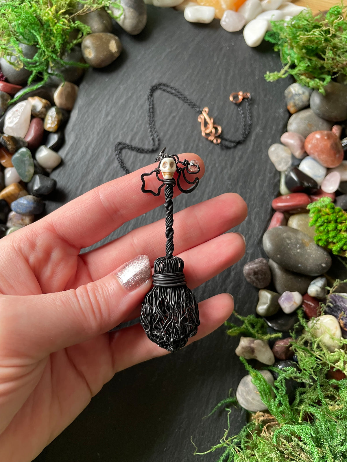 A hand holding a black broom pendant with a small skull bead, shown against a dark slate background with decorative stones and moss.