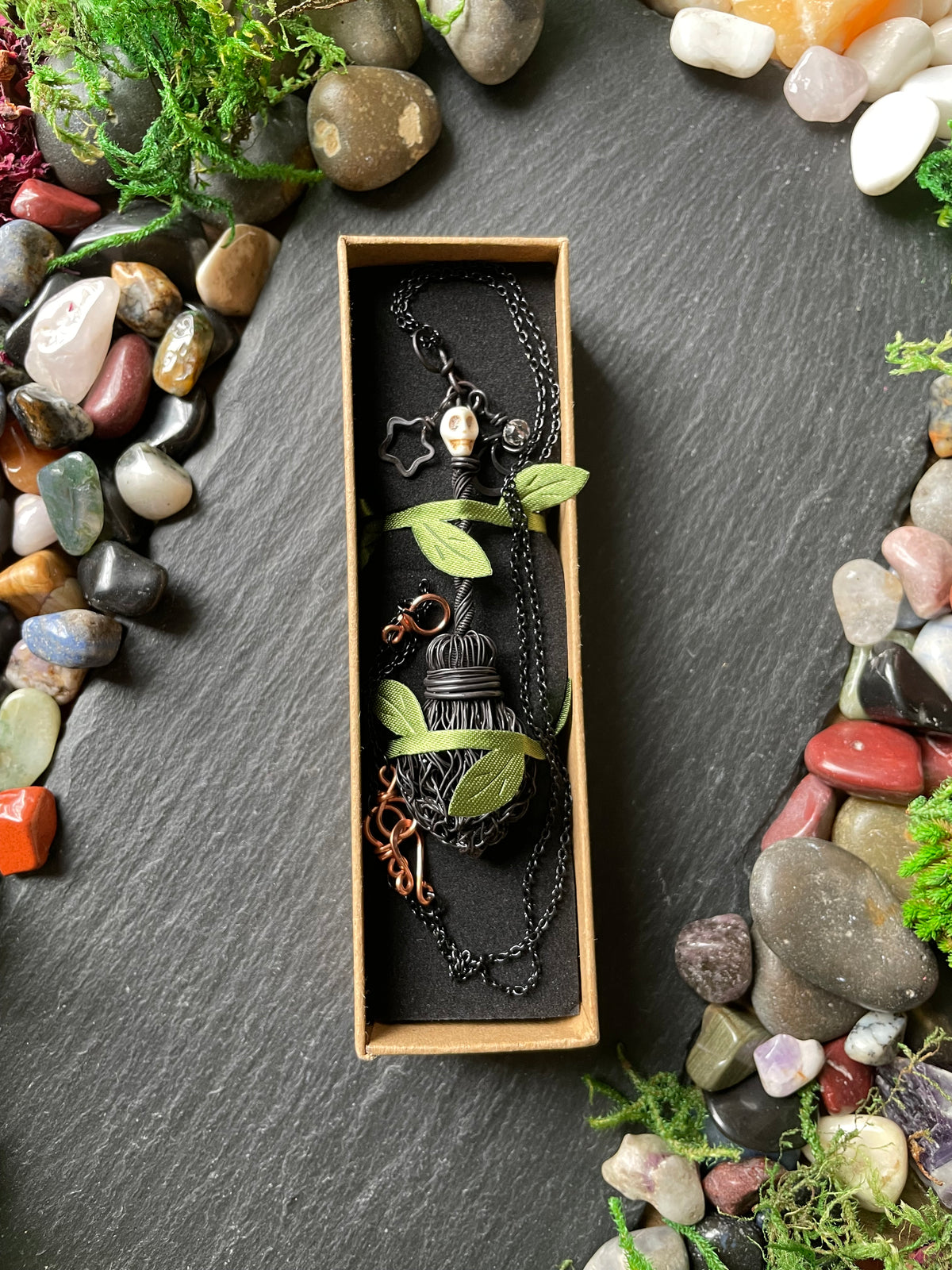 A black metal necklace chain with a handmade broom pendant attached, displayed inside a rectangular cardboard box on a dark slate background surrounded by decorative stones and moss.