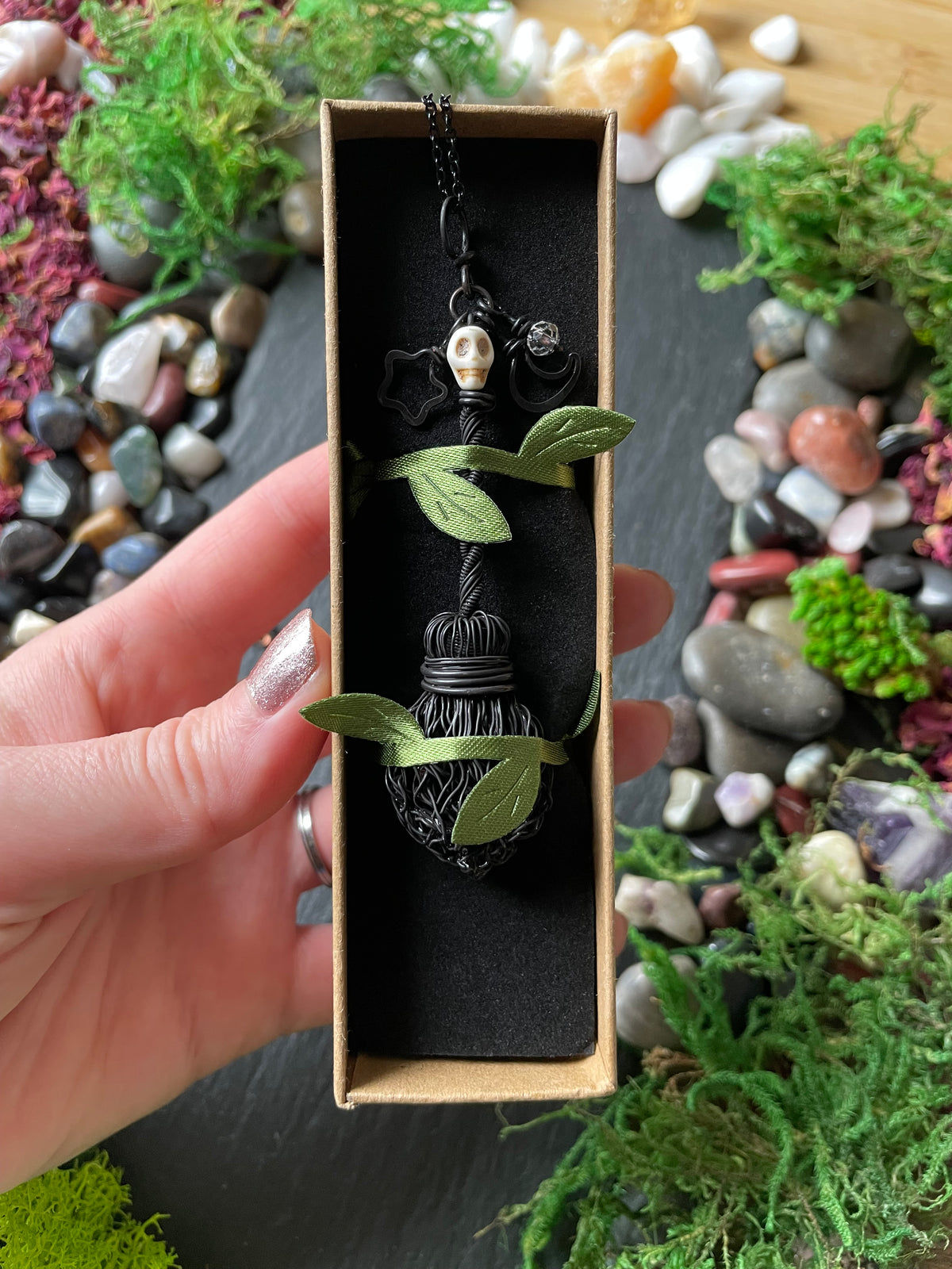 A hand holds a box containing a black gothic-style broom pendant necklace with a skull bead. The background is a styled flat-lay of moss, pebbles, and crystals on a dark slate surface.