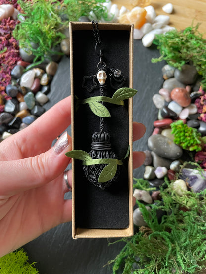 A hand holds a box containing a black gothic-style broom pendant necklace with a skull bead. The background is a styled flat-lay of moss, pebbles, and crystals on a dark slate surface.