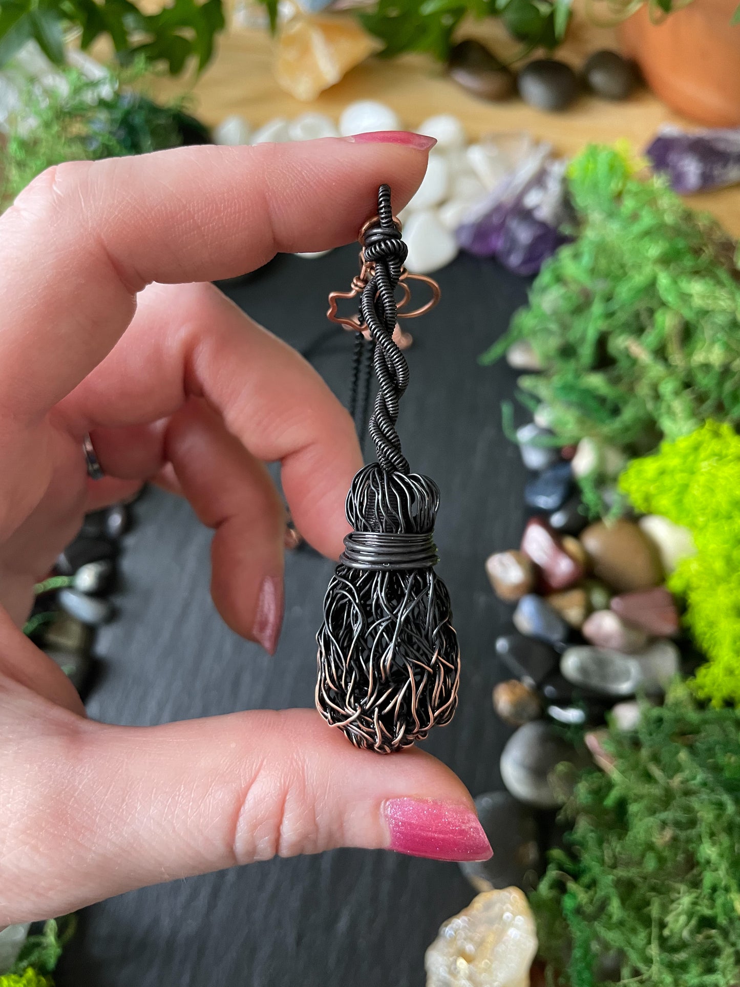 A hand holds a black and copper wire-wrapped pendant against a styled background of green moss, small stones, and crystals.