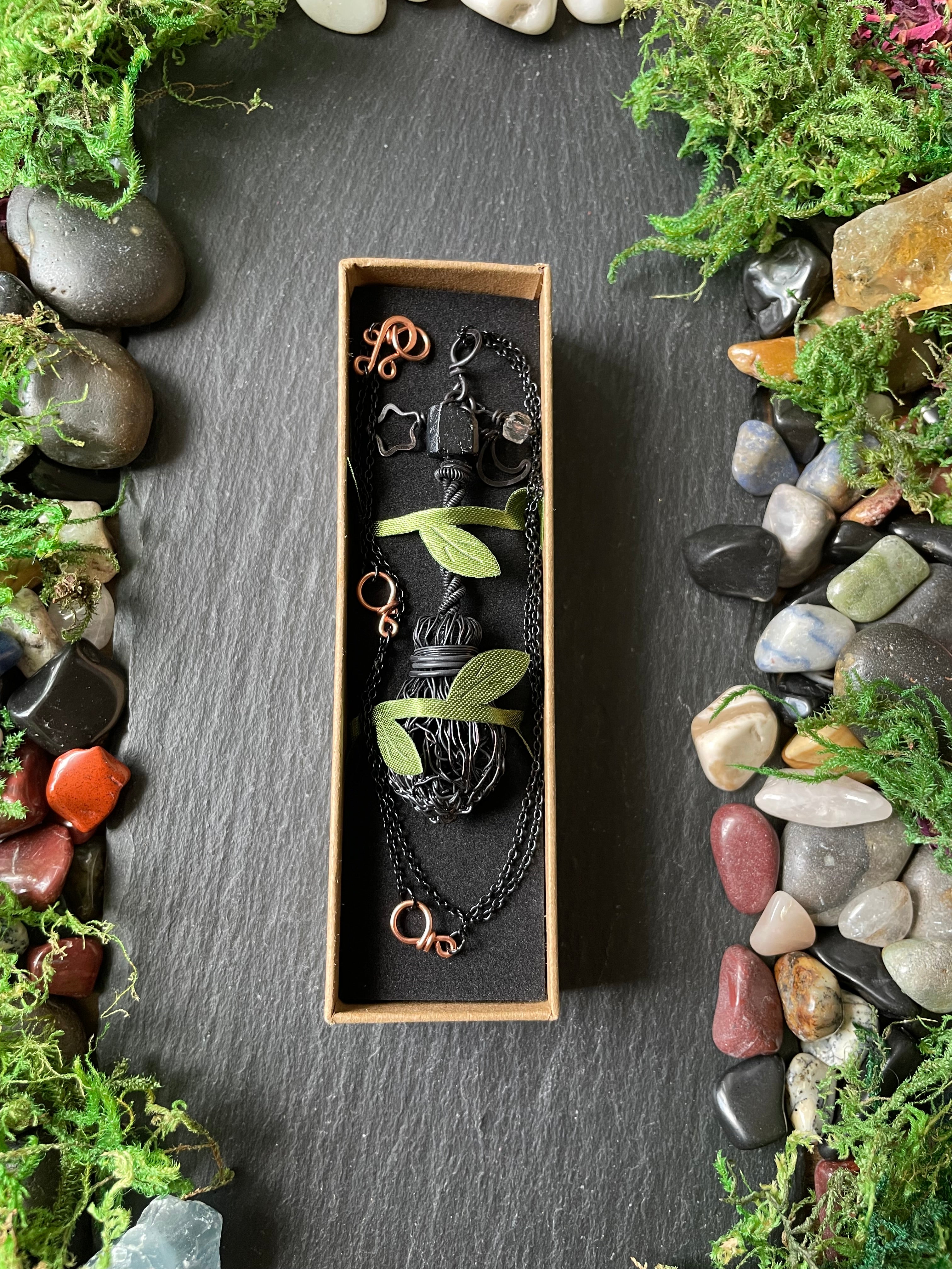 A black mini broom pendant in box on a dark slate surface surrounded by moss and stones.