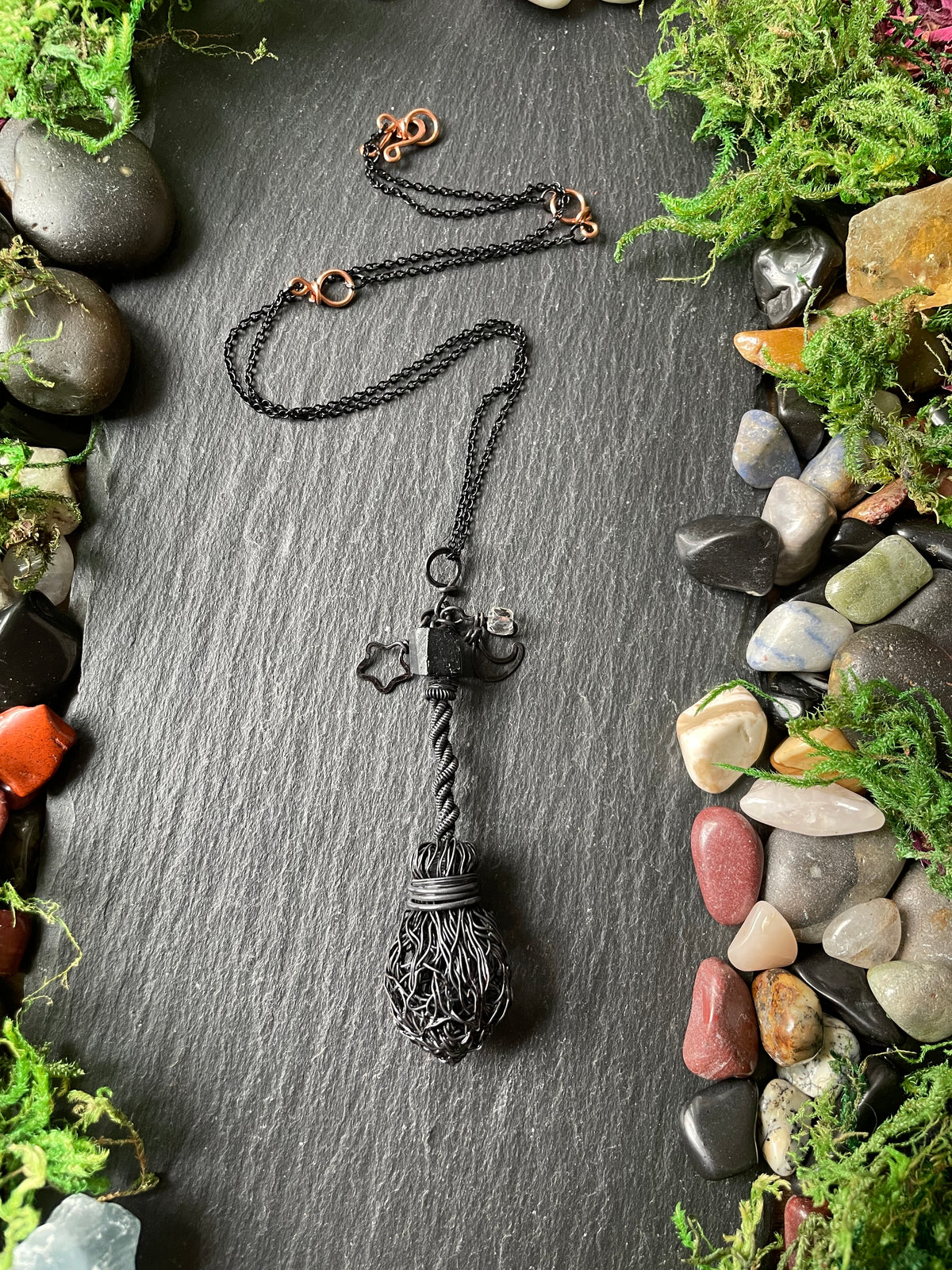 A black broom pendant displayed on a dark slate background surrounded by green moss and smooth stones.