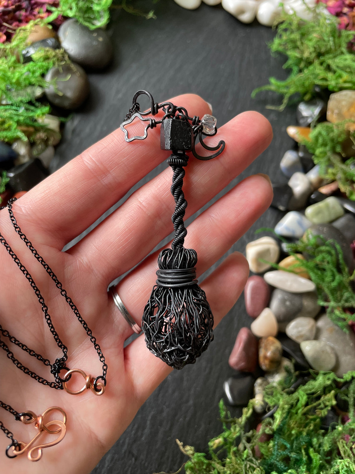 A hand holding a dark broom necklace set against a background of moss and small stones.