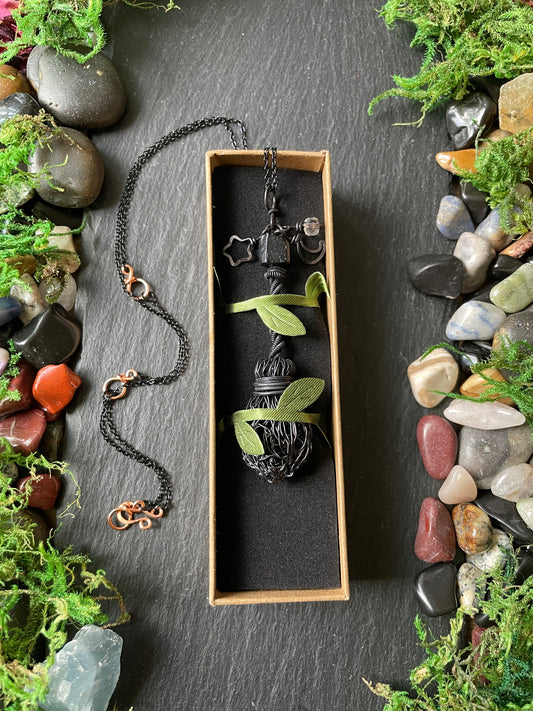 A black mini handmade broom pendant, displayed in a rectangular box with green leaves on a dark slate surface surrounded by moss and stones.