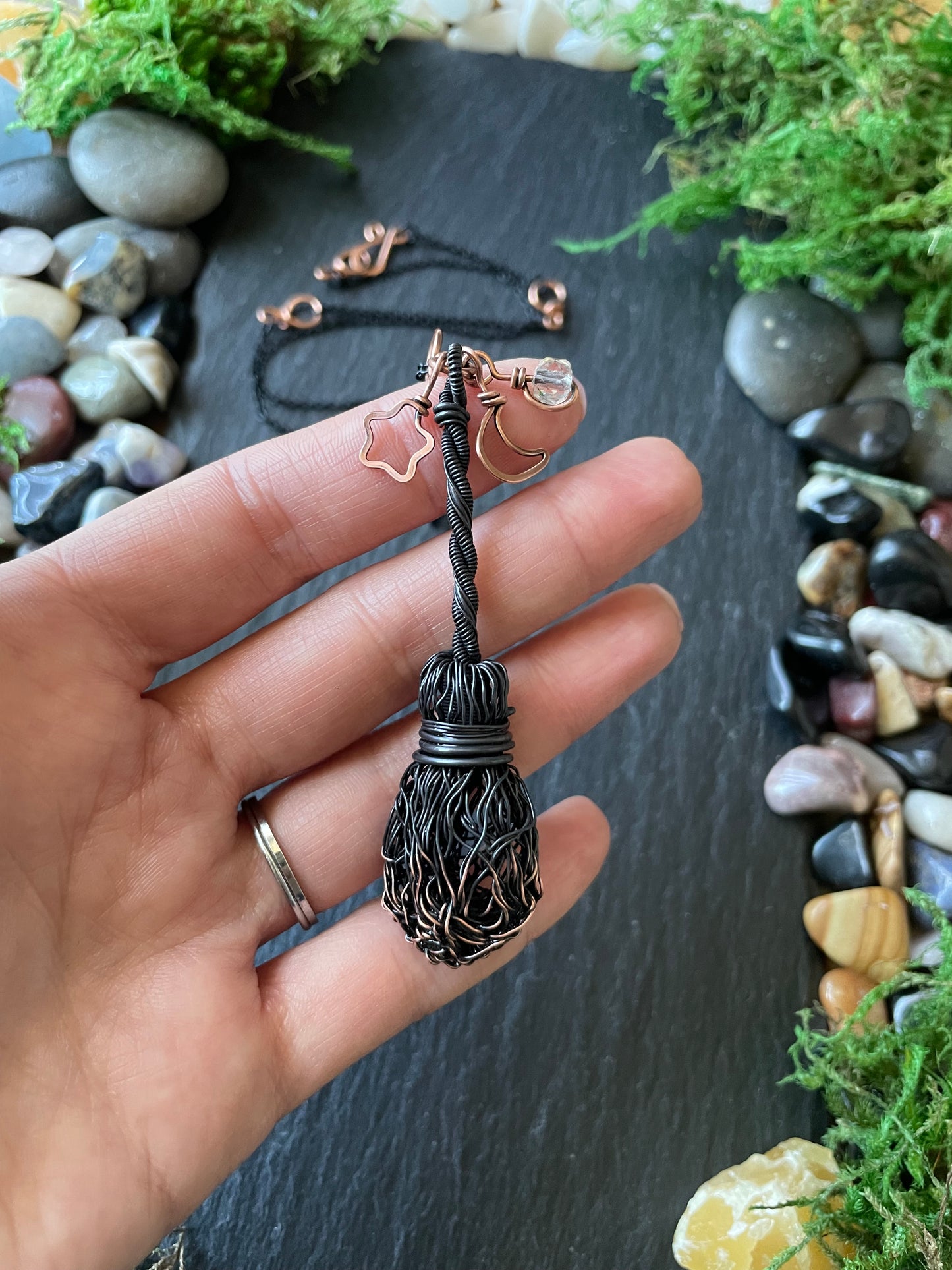 Hand holding a decorative broom pendant with a natural stone and moss background