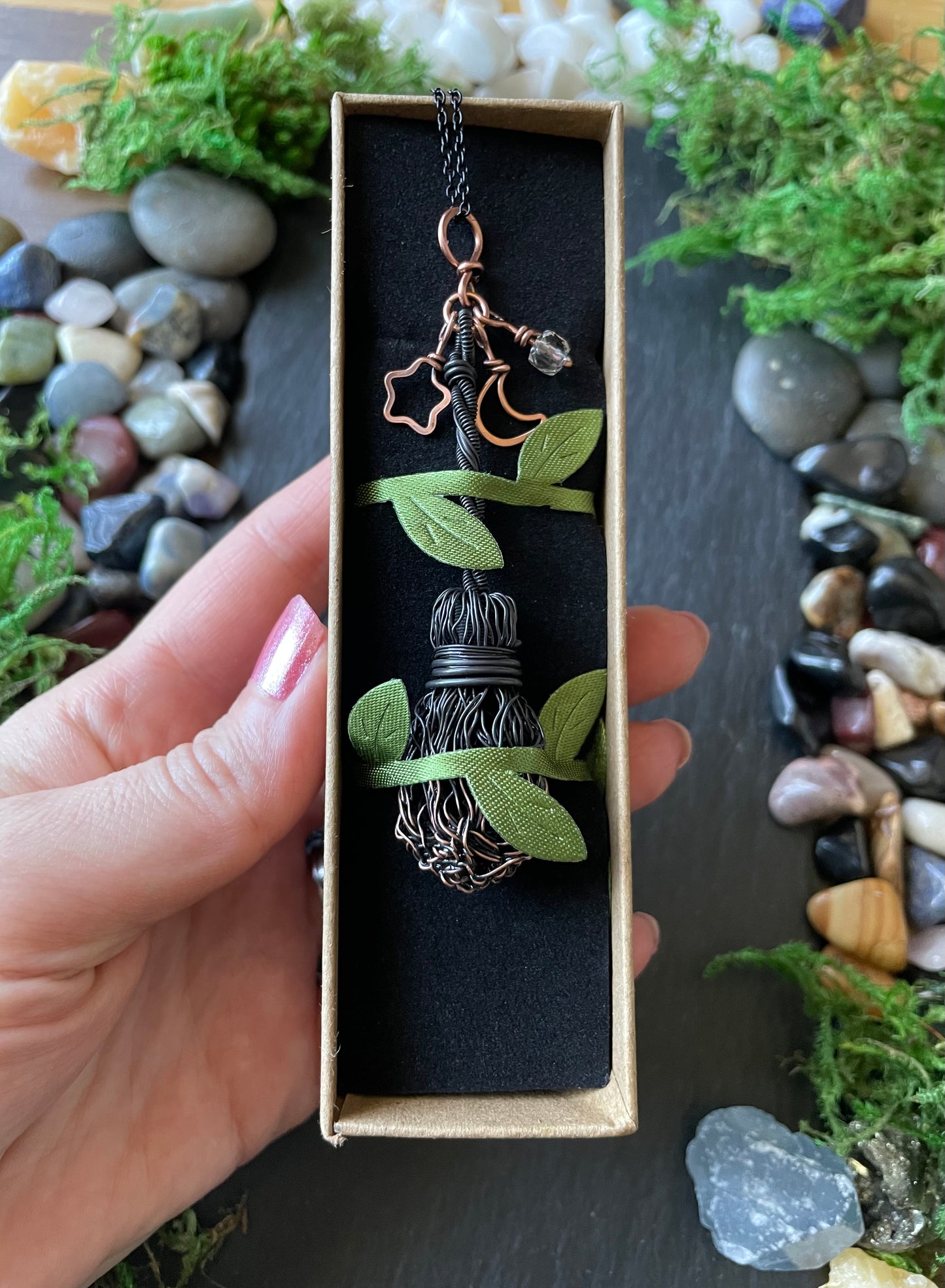 Hand holding a decorative pendant with green leaves and a star charm in a box, surrounded by stones and plants.