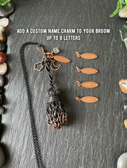 A custom copper name charm for a broom, shown as a black wire-wrapped broom with a copper name plate , alongside four example copper name plates engraved with names. The image is set on a dark slate background with decorative stones and features white text overlay that reads 'ADD A CUSTOM NAME CHARM TO YOUR BROOM UP TO 8 LETTERS'.