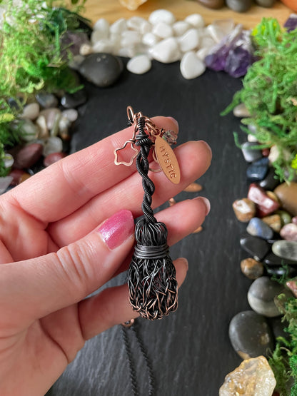A hand holding a handmade metal broom featuring an oval copper charm engraved with 'MYSTIC'.