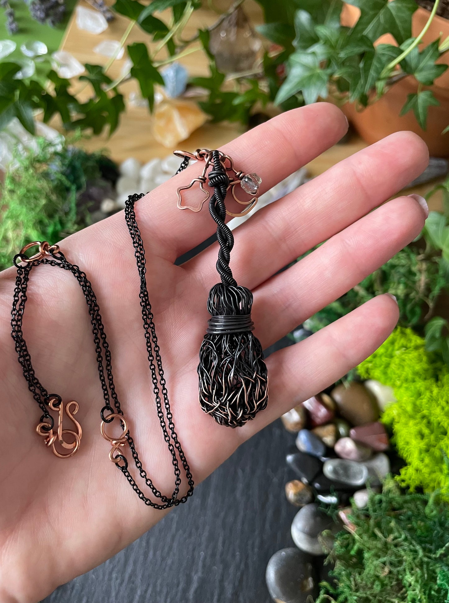A handmade dark metal broom necklace held in a person's hand against a background of green plants and moss.