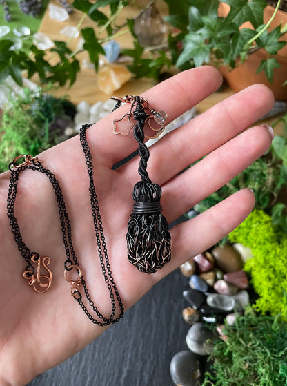 A handmade dark metal broom necklace held in a person's hand against a background of green plants and moss.
