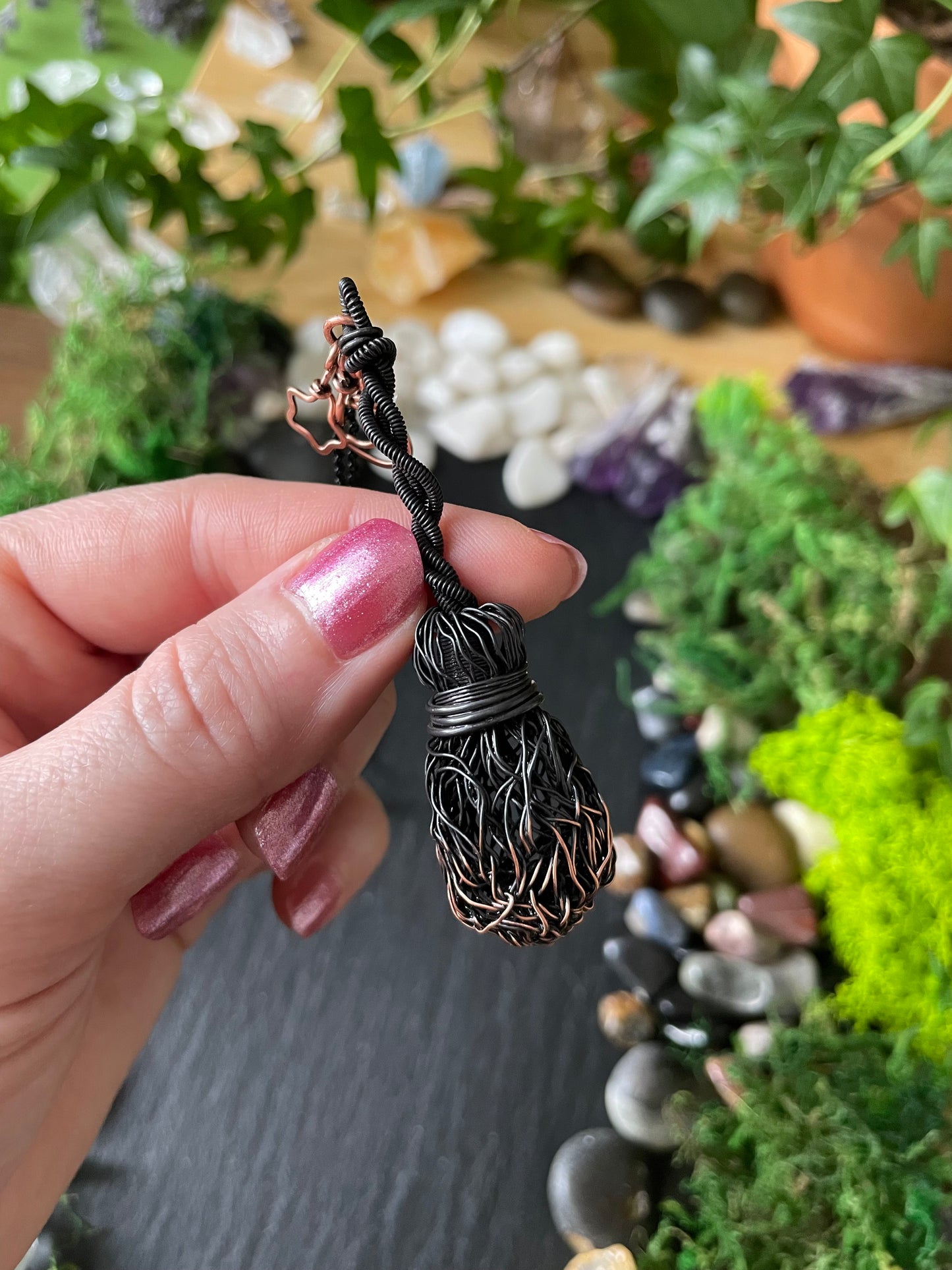 A hand holds a black wire broom pendant against a blurred background of plants, moss, and stones.