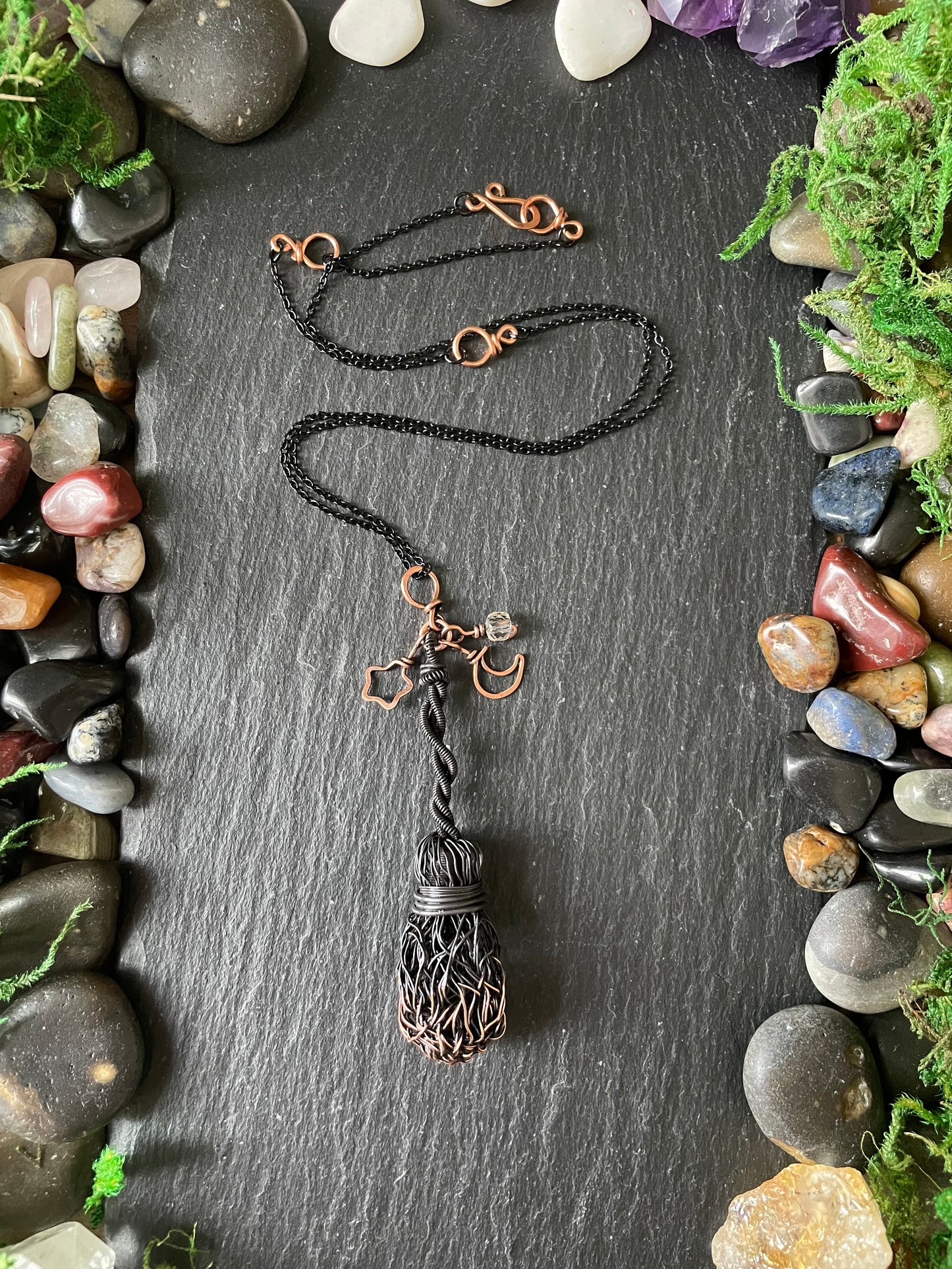A handmade black broom pendant displayed on a dark slate background surrounded by decorative stones and greenery.
