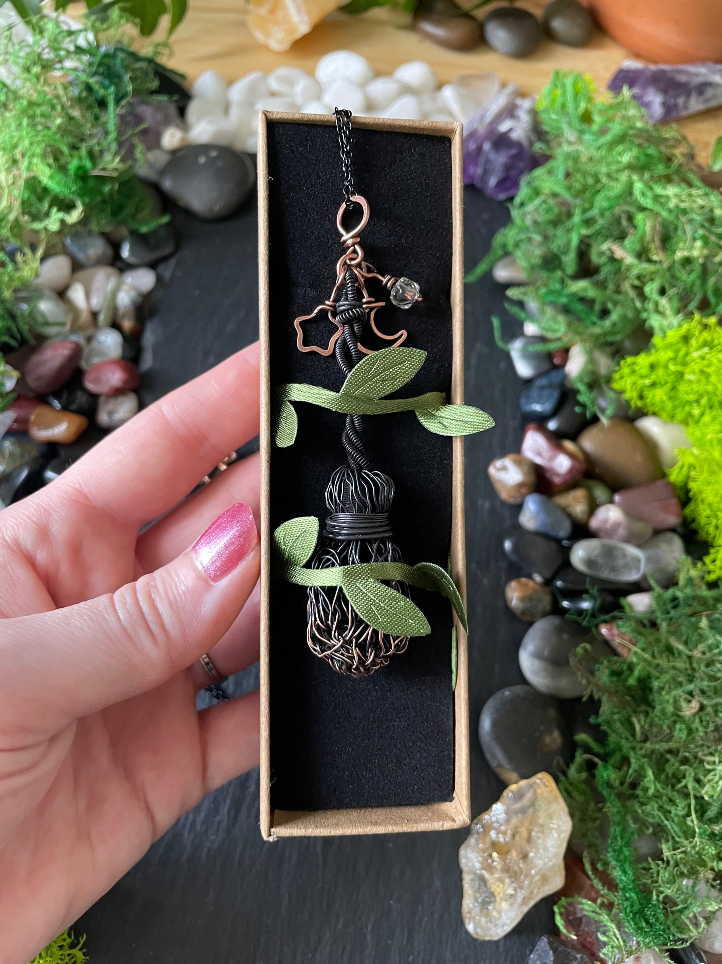 A hand holds a small rectangular jewelry box containing a handmade wire-wrapped pendant necklace with green leaf-shaped accents and a copper star charm, set against a background of moss, stones, and crystals.