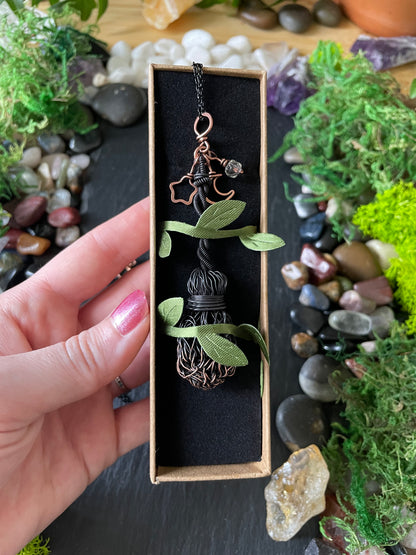 A hand holds a small rectangular jewelry box containing a handmade wire-wrapped pendant necklace with green leaf-shaped accents and a copper star charm, set against a background of moss, stones, and crystals.