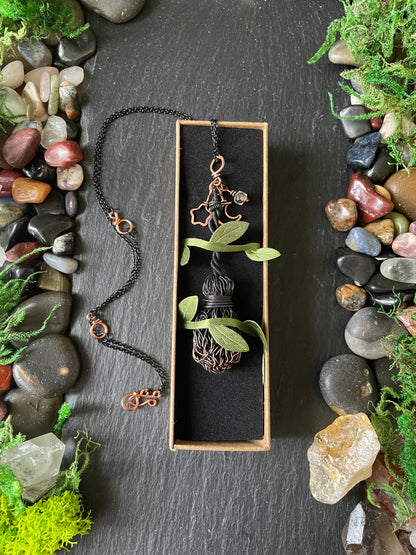 A copper broom pendant, displayed in a small box on a black background, surrounded by stones, moss, and greenery.