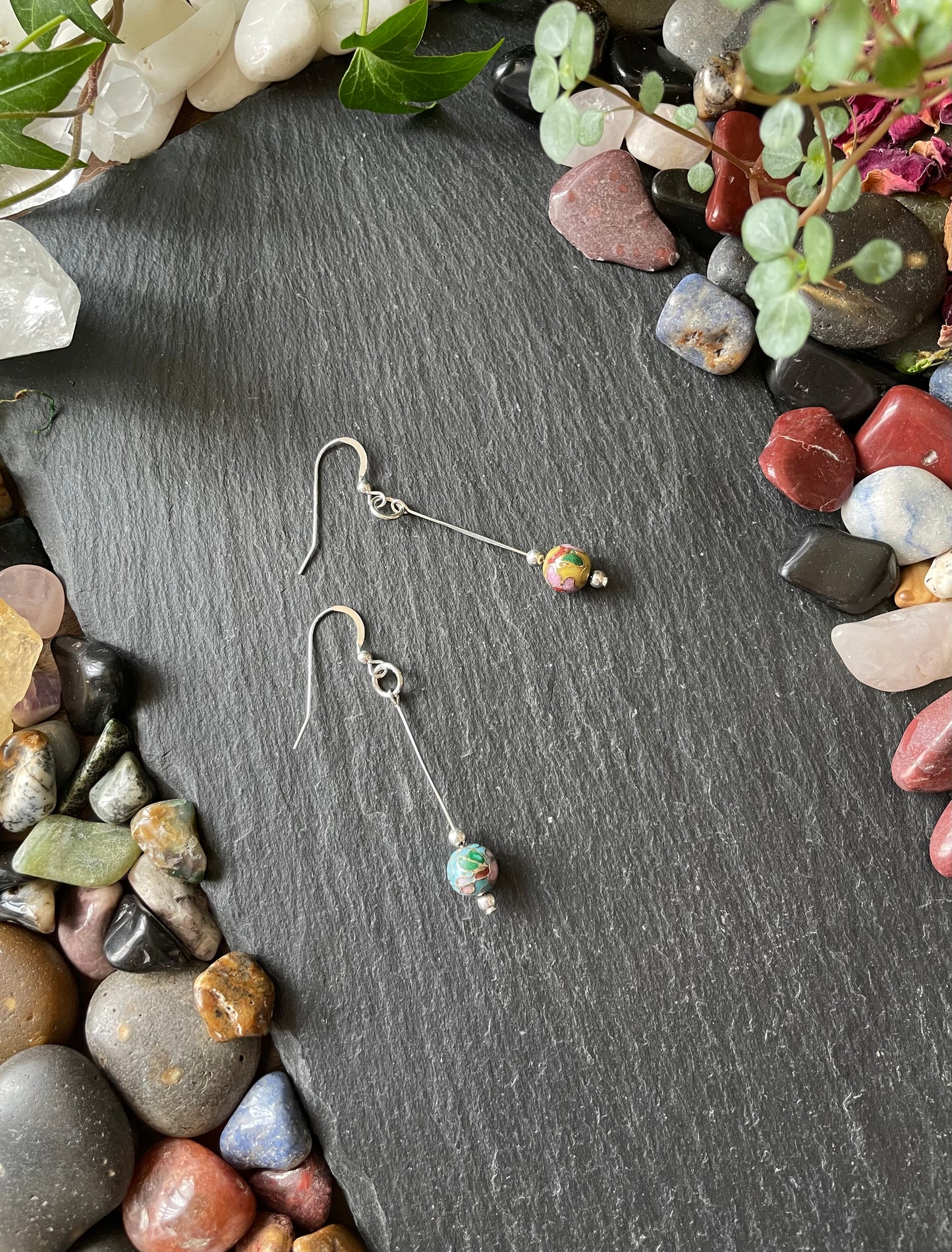 Pair of silver earrings with beads on a dark stone surface surrounded by small rocks and greenery
