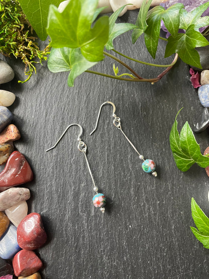 Silver earrings with colorful blue beads on a dark surface with green leaves and stones.