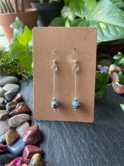 Pair of silver earrings with flower beads on a brown card, set against a natural stone and plant background.