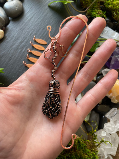 Hand holding a copper necklace with a decorative pendant against a natural stone and moss background