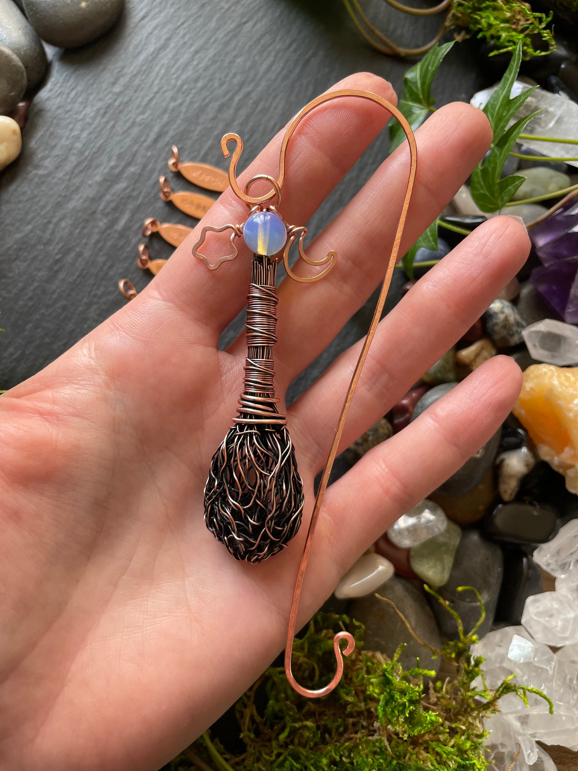 Hand holding a copper wire-wrapped pendant with a natural stone against a background of stones and crystals.