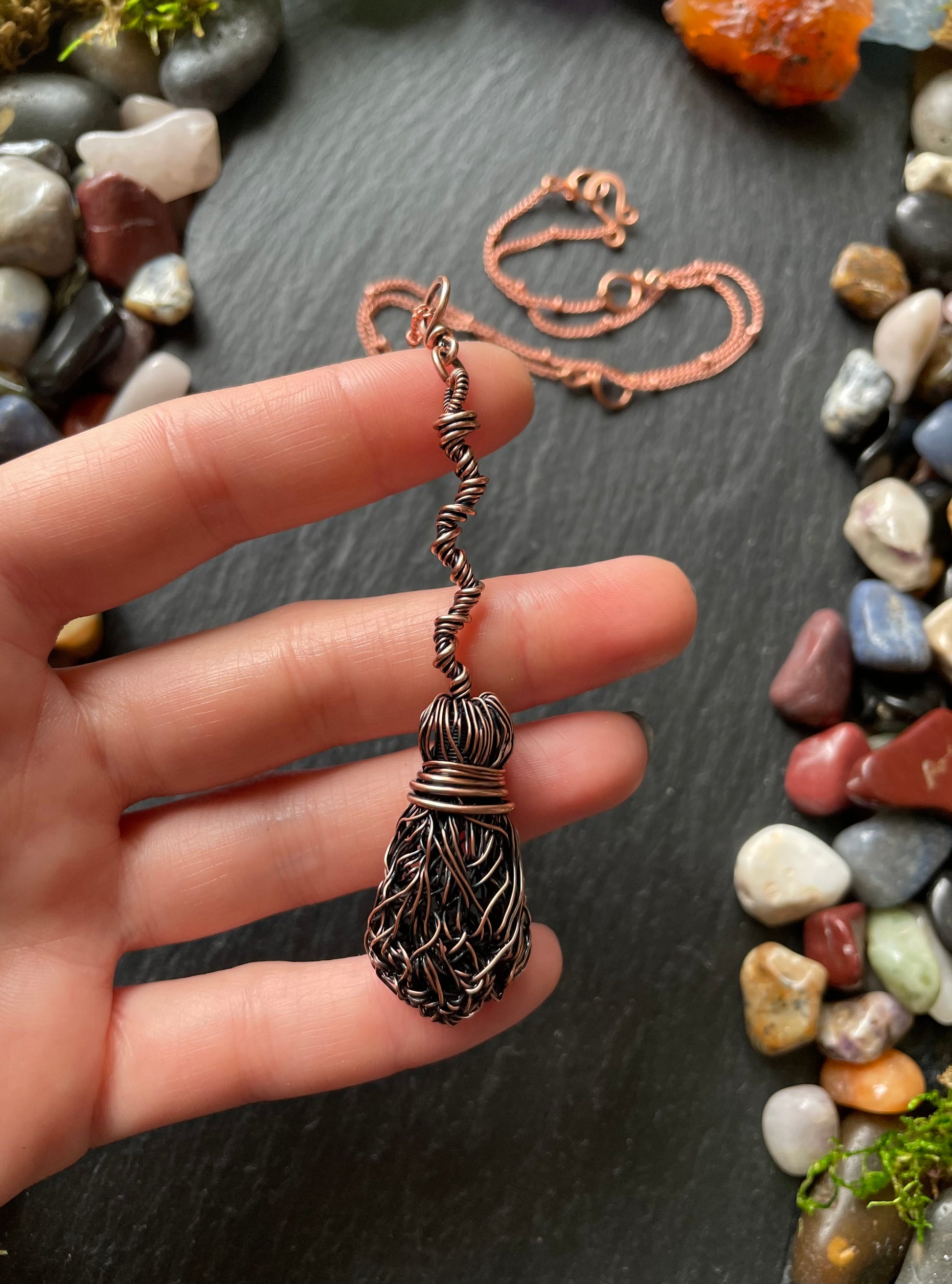 Hand holding a copper wire-wrapped pendant with pebbles in the background