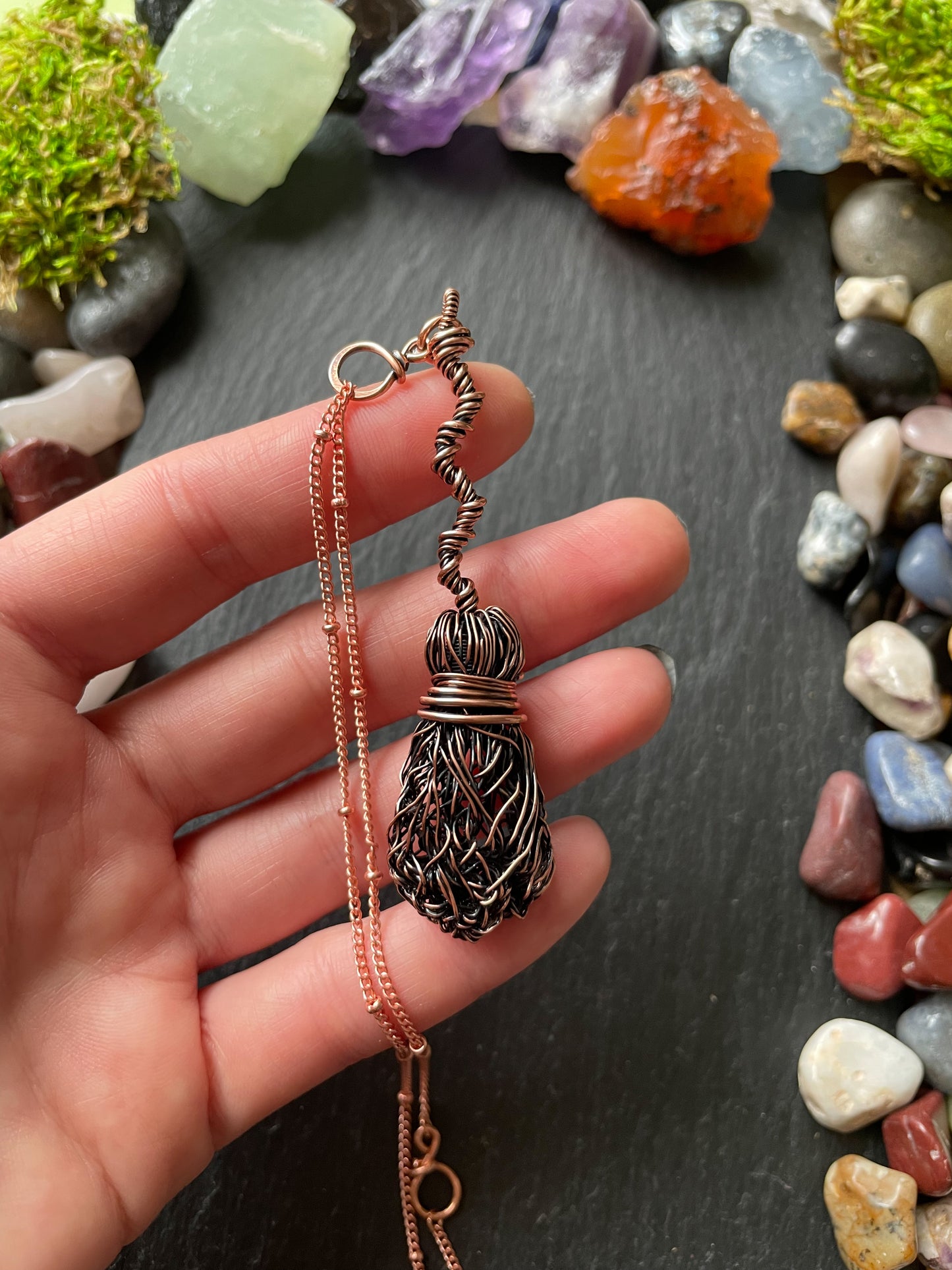 Hand holding a copper necklace with a decorative pendant against a background of colorful stones.