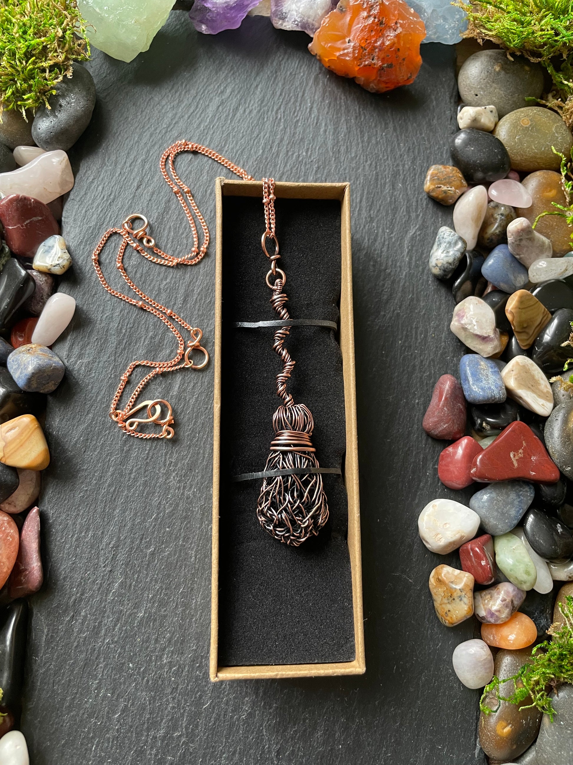 Necklace with a broom pendant in a box surrounded by stones and crystals on a dark surface