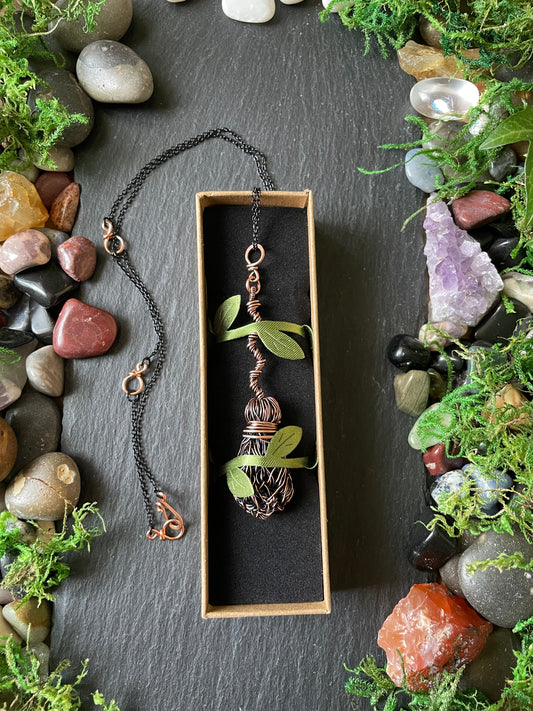 A handmade copper pendant necklace displayed in a small box on a dark slate background, surrounded by decorative stones and moss.