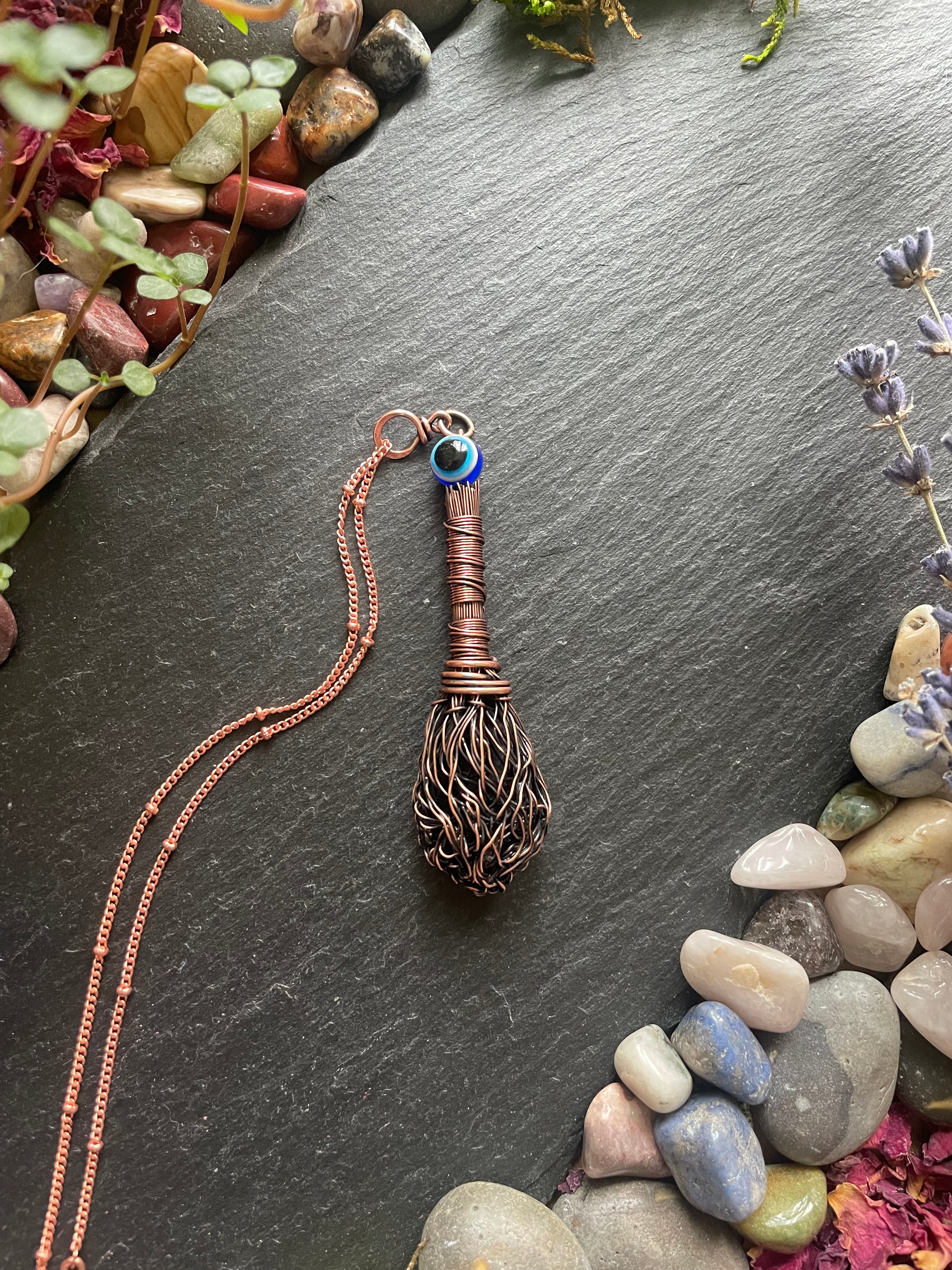 Broom necklace with an evil eye bead on a dark stone surface with pebbles and plants.