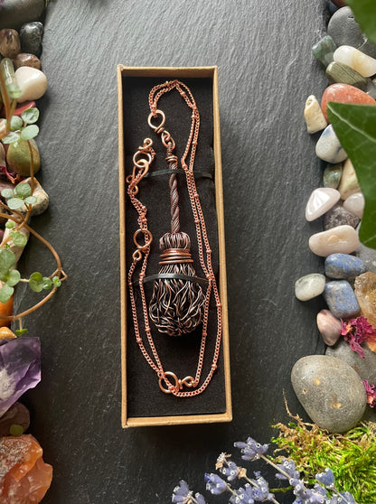 Copper broom necklace in a box on a dark surface with stones and plants around