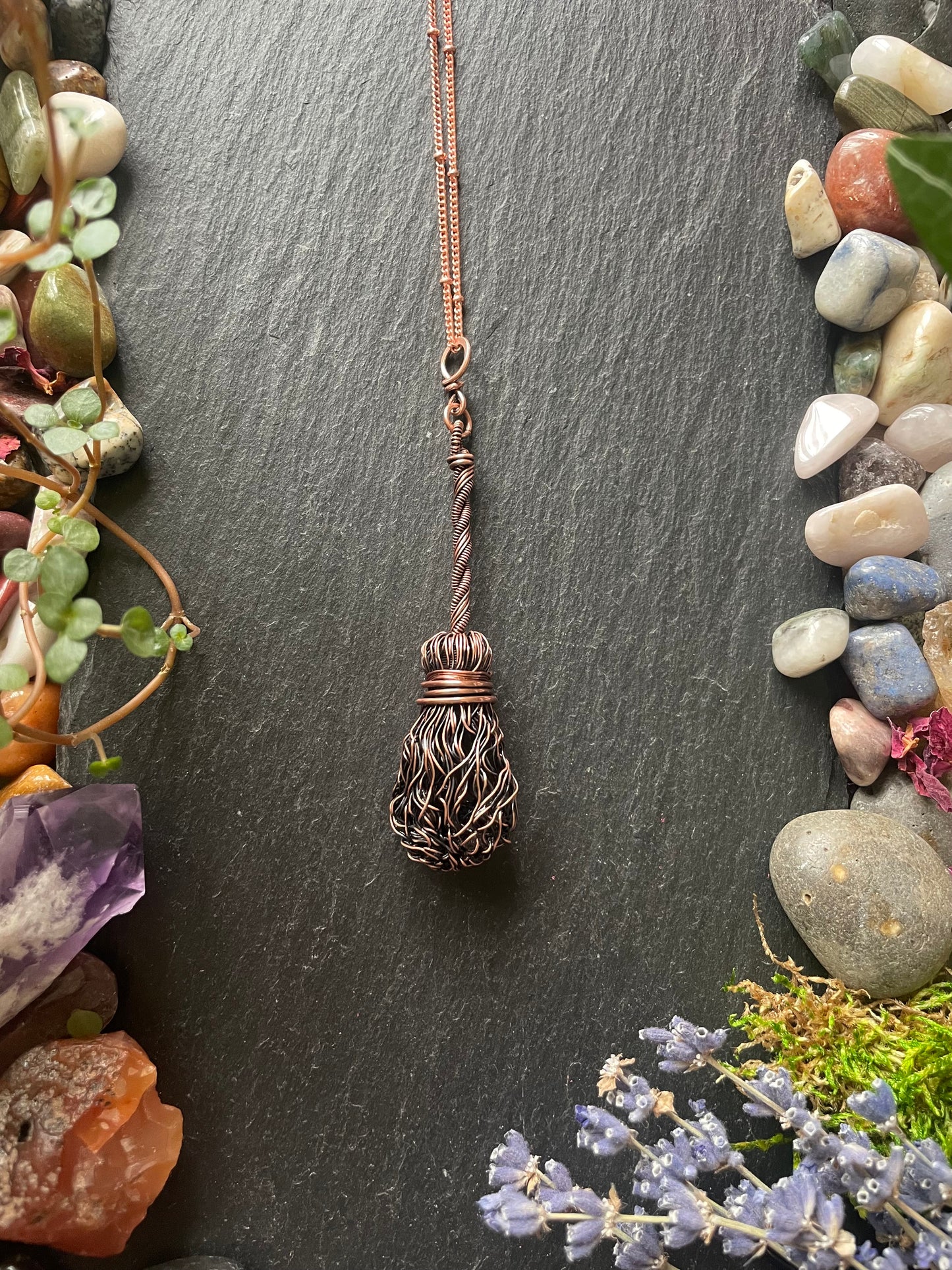 Copper witch broom necklace on a dark slate background with stones and plants.