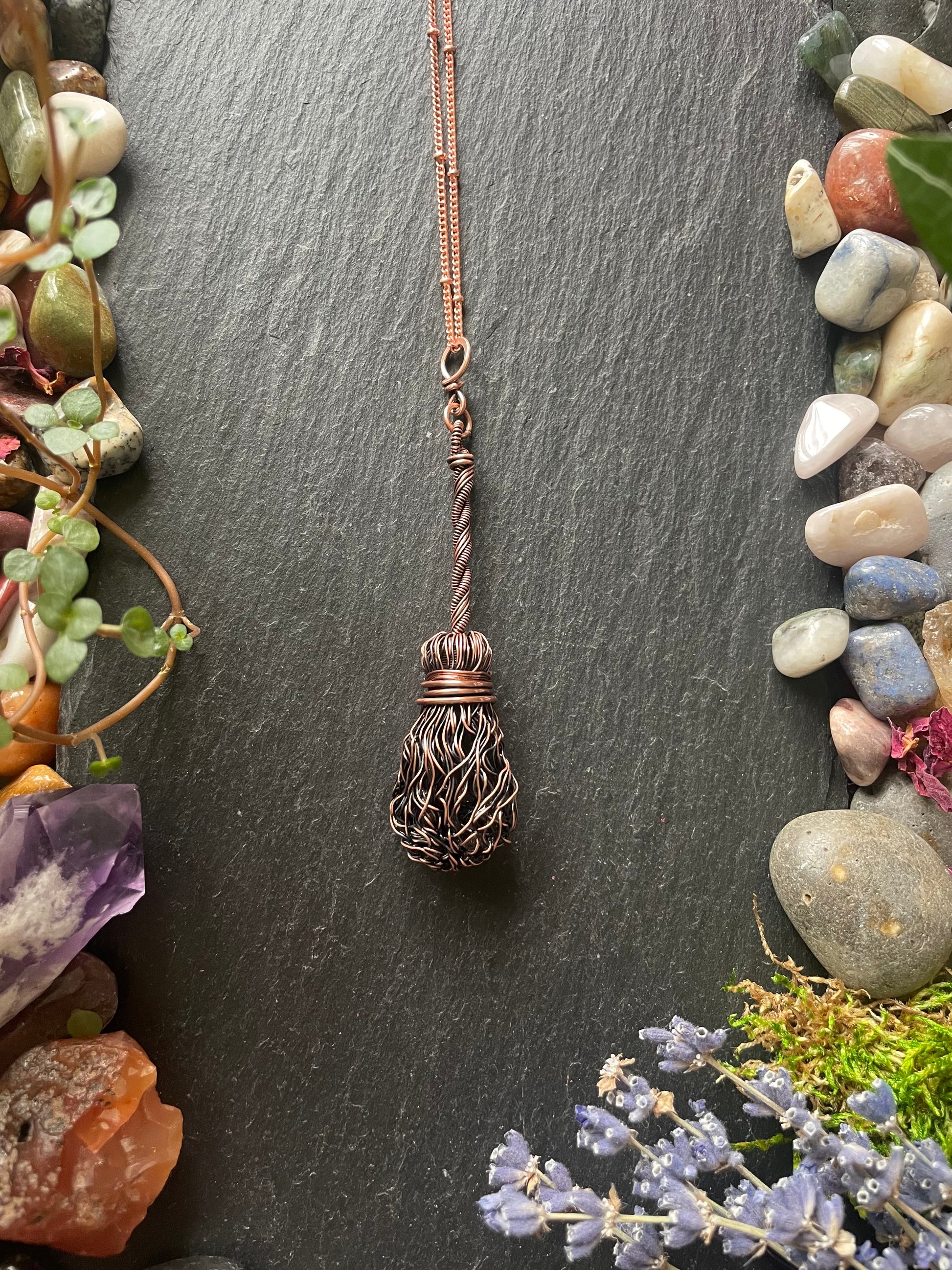 Copper witch broom necklace on a dark slate background with stones and plants.