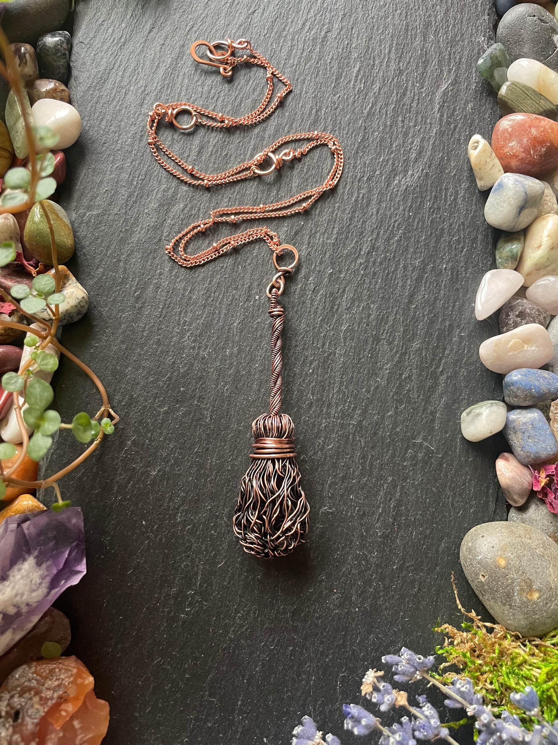 Decorative witch's broom pendant on a dark stone surface surrounded by stones and plants.