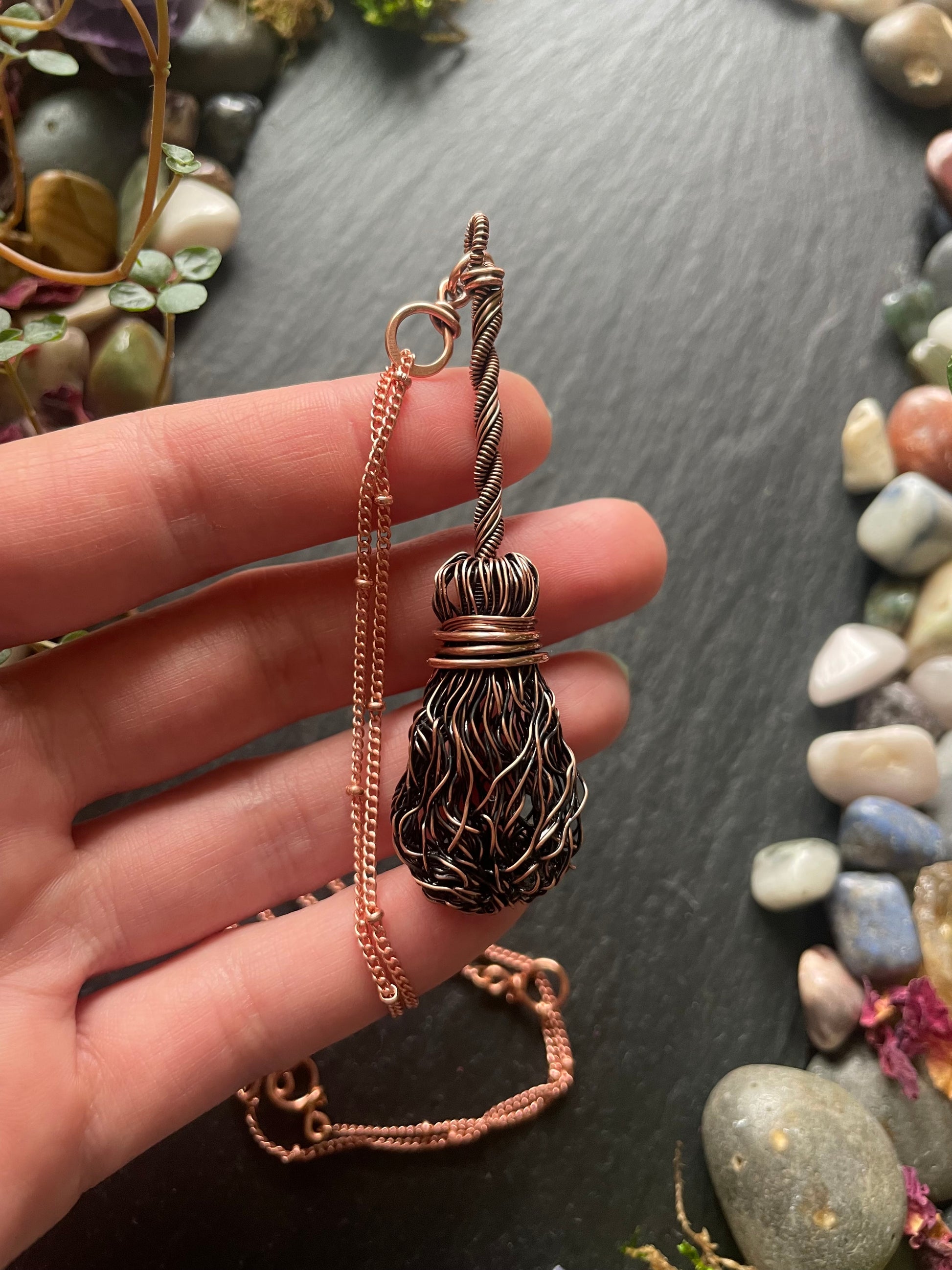 Hand holding a copper witch's broom necklace on a dark surface with pebbles.