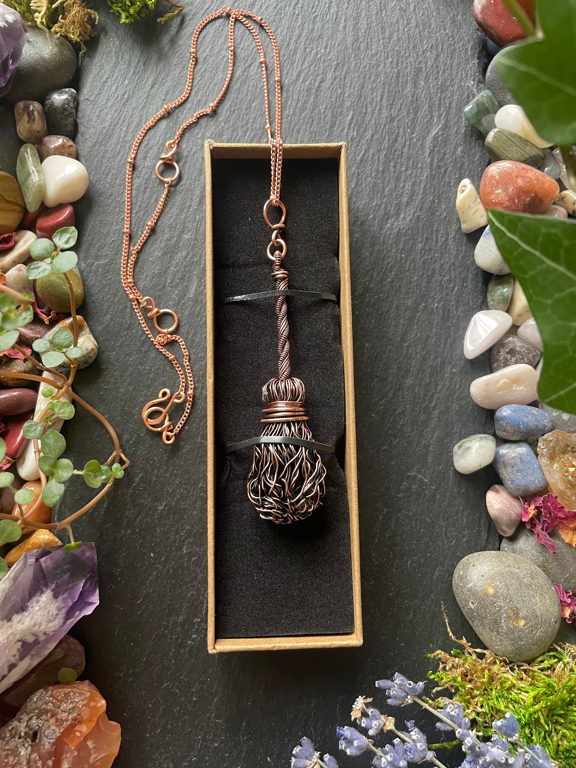 Copper necklace with a broom pendant in a box, surrounded by stones and plants
