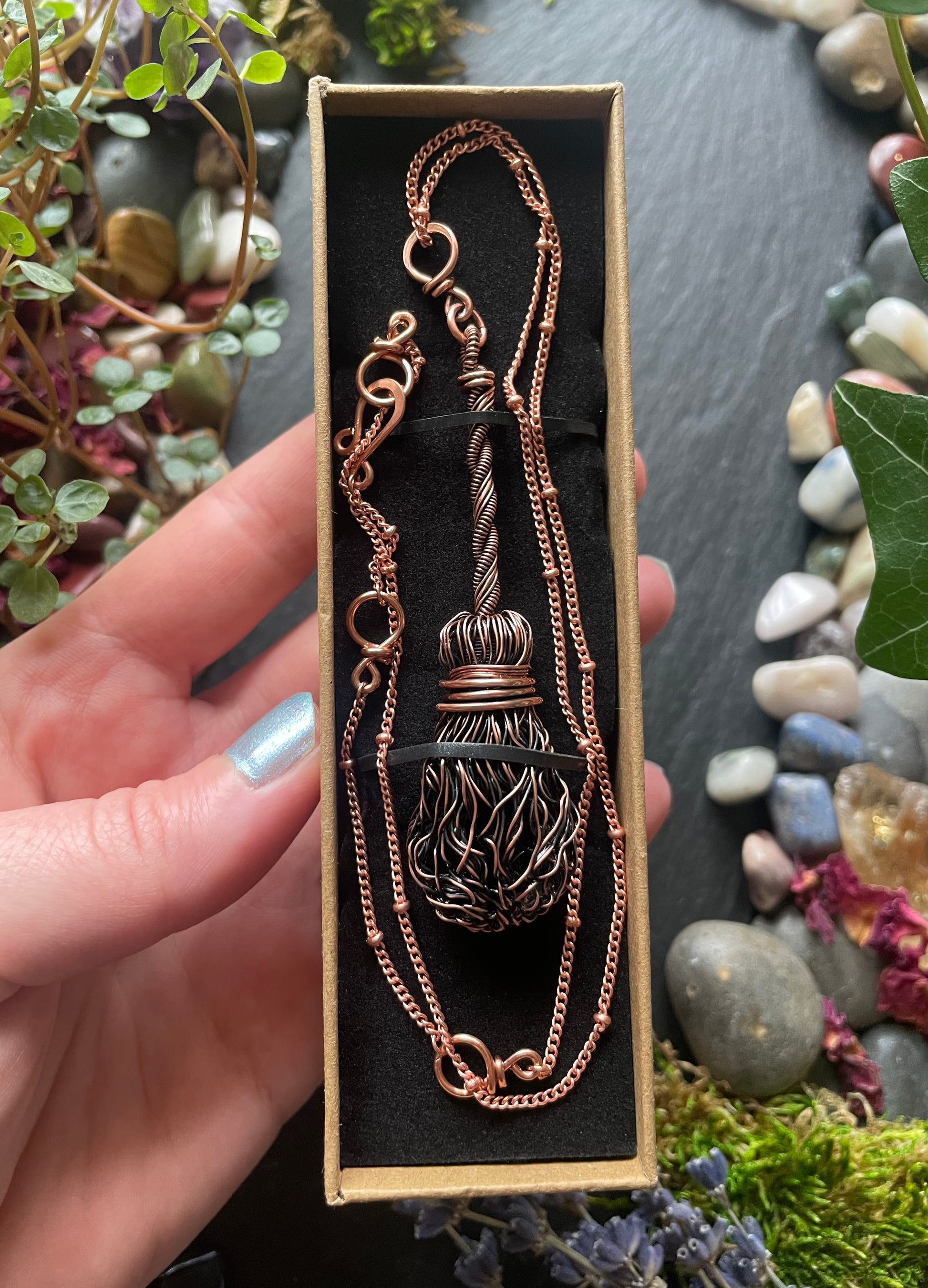 Copper necklace with broom pendant in a box held by a hand with a natural background of stones and plants.